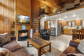 A spacious living area with a fireplace, a wooden staircase, a kitchen, and a dining area.