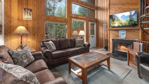 Ein gemütliches Blockhaus-Interieur mit Kamin, Ledersofas, einem Couchtisch und großen Fenstern mit Blick auf den Wald.