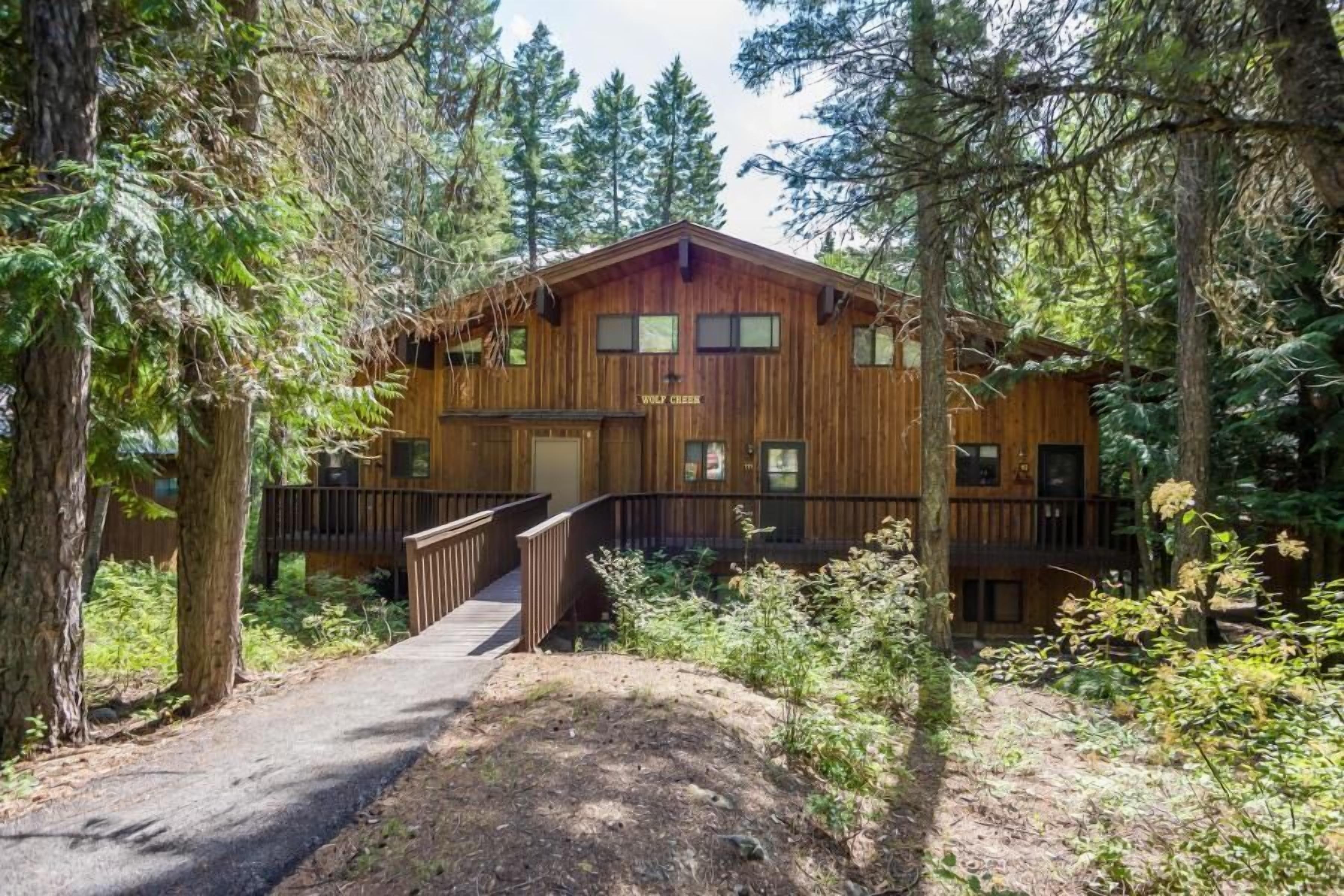 A wooden cabin with a deck and a covered walkway, surrounded by dense forest.