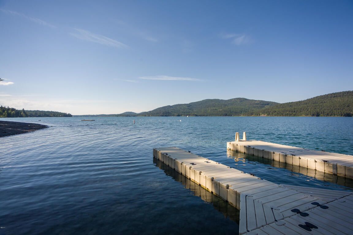 A wooden dock extending into calm water with a clear sky and forested hills in the background.