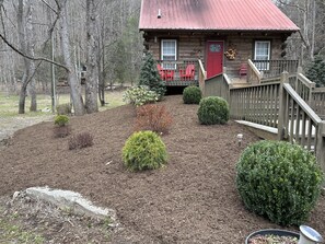 A rustic cabin with a red door, a wooden deck, and a landscaped garden with mulch and shrubs. - BRAND NEW JACUZZI HOT TUB Real Log Cabin, Beautiful Rushing Trout Stream, Wifi. (Deep Gap)
