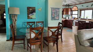 Un coin repas avec une table ronde en verre et des chaises en bois, une cuisine avec des meubles en bois et une fenêtre, et un plafond avec des poutres apparentes et des luminaires suspendus.