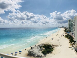 A beach with clear blue water, sandy shore, and a few people. There are buildings on the right side of the image.