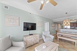 A spacious living room with a flat-screen TV, a wooden cabinet, a white armchair, and a dining area with benches. - Seawind 1703 by Liquid Life (Gulf Shores)