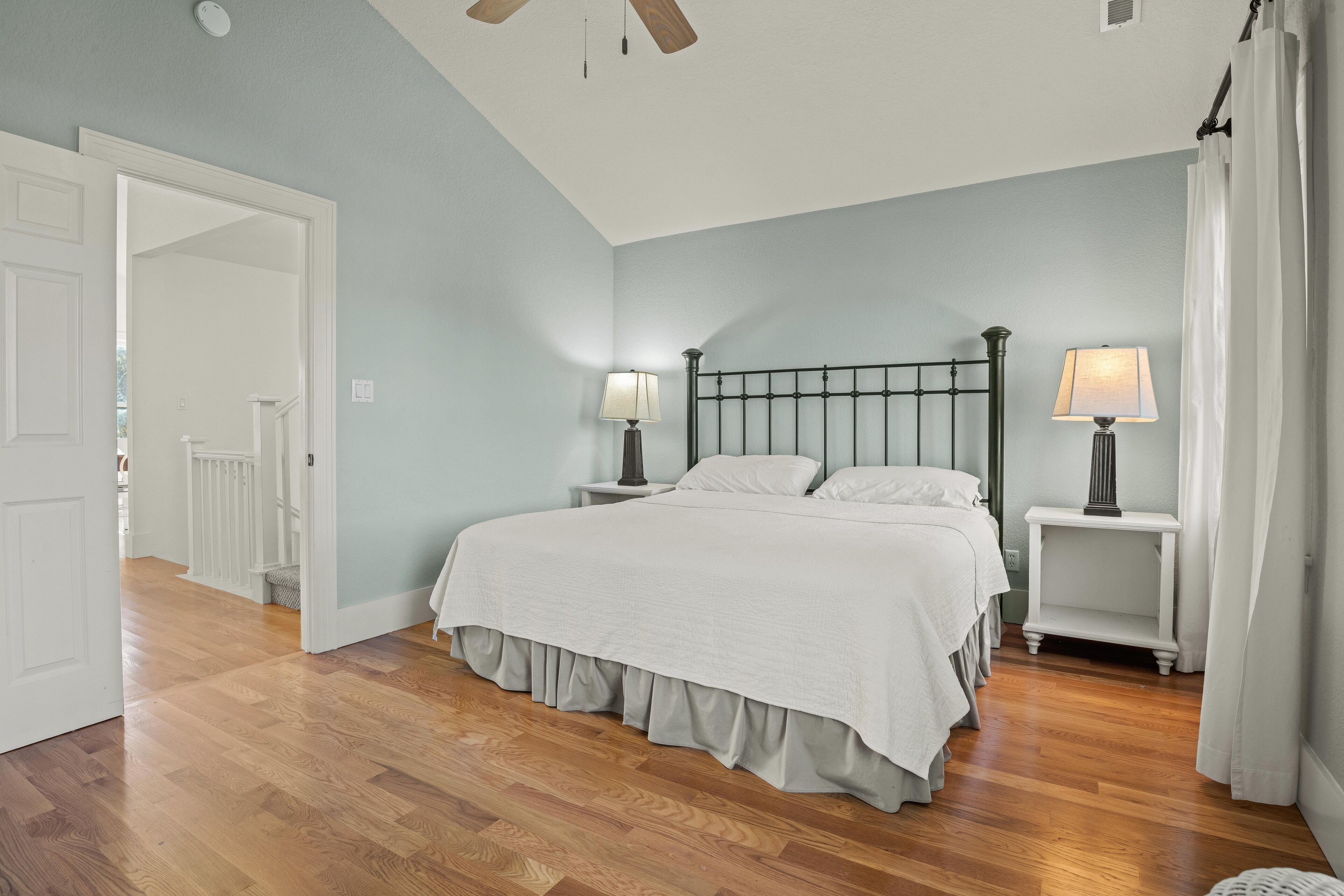 Une chambre à coucher comprenant un lit, deux tables de chevet avec des lampes, un ventilateur de plafond et une porte donnant sur une autre pièce.
