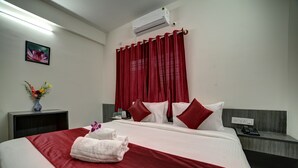 Una habitación de hotel con una cama grande, cortinas rojas, aire acondicionado montado en la pared, una mesita de noche con teléfono y un jarrón con flores.