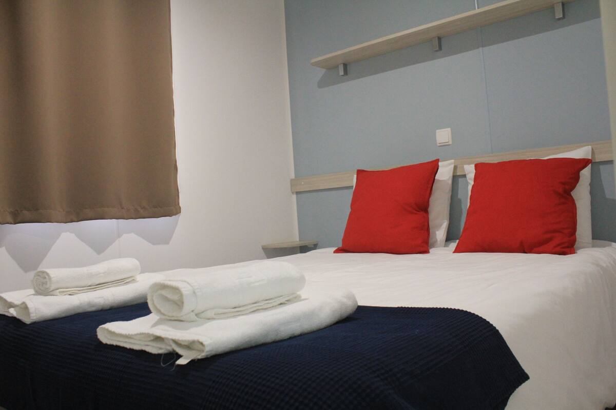 Una cama bien hecha con sábanas blancas, dos almohadas rojas, una manta azul y una cortina marrón.