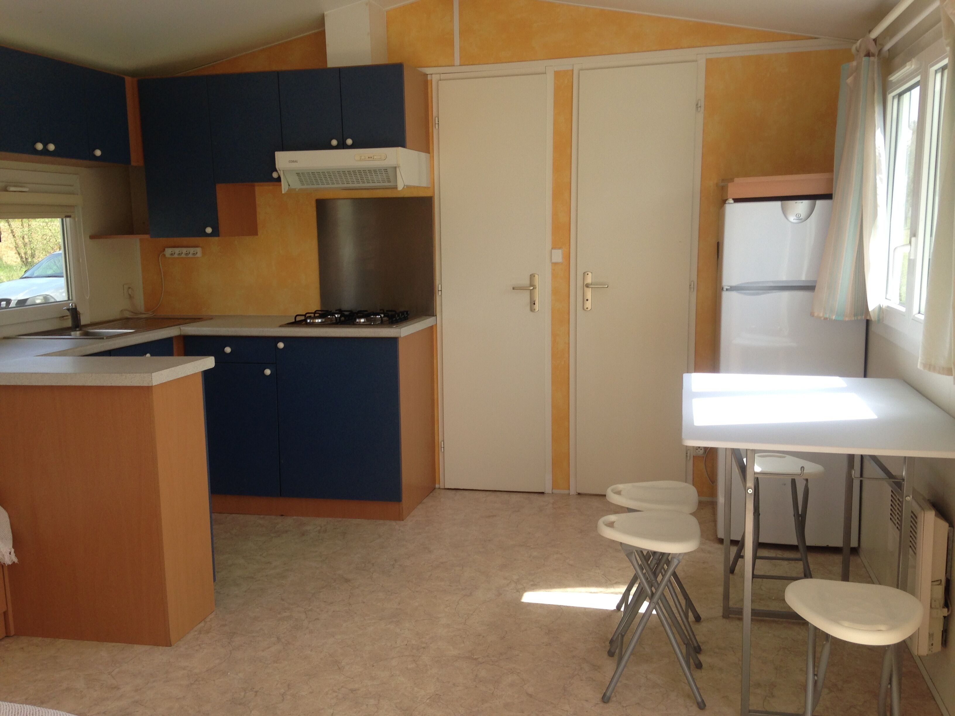 A compact kitchen with a gas stove, a built-in oven, and a refrigerator. There are two stools and a small table.