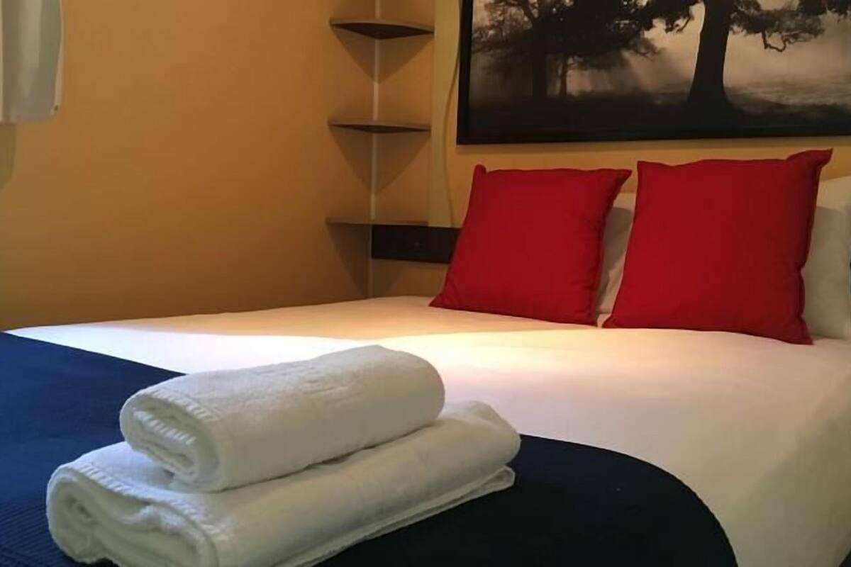 Una cama bien hecha con almohadas blancas y rojas, una toalla blanca doblada y una colcha azul.