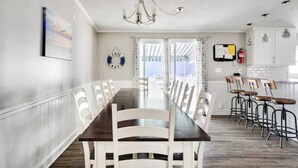 Un espace repas avec une longue table, des chaises blanches, un lustre et une vue dégagée sur l’extérieur grâce à de grandes fenêtres.