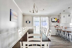 A dining area with a long table, white chairs, a chandelier, and a view through large windows.