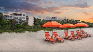 Ein Strand mit roten Sonnenschirmen und Liegestühlen, ein mehrstöckiges Gebäude und ein Sonnenuntergangshimmel.