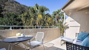 En balkon med et rundt bord, en stol og en sofa. Der er krukker med planter og udsigt til træer og bakker.