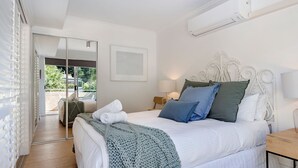 Een slaapkamer met een bed, nachtkastje, lamp, een wit hoofdeinde en een airconditioning.