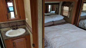 En kompakt autocamper indretning med badeværelse, der indeholder vask og spejl, en seng med puder og et vindue med gardiner.