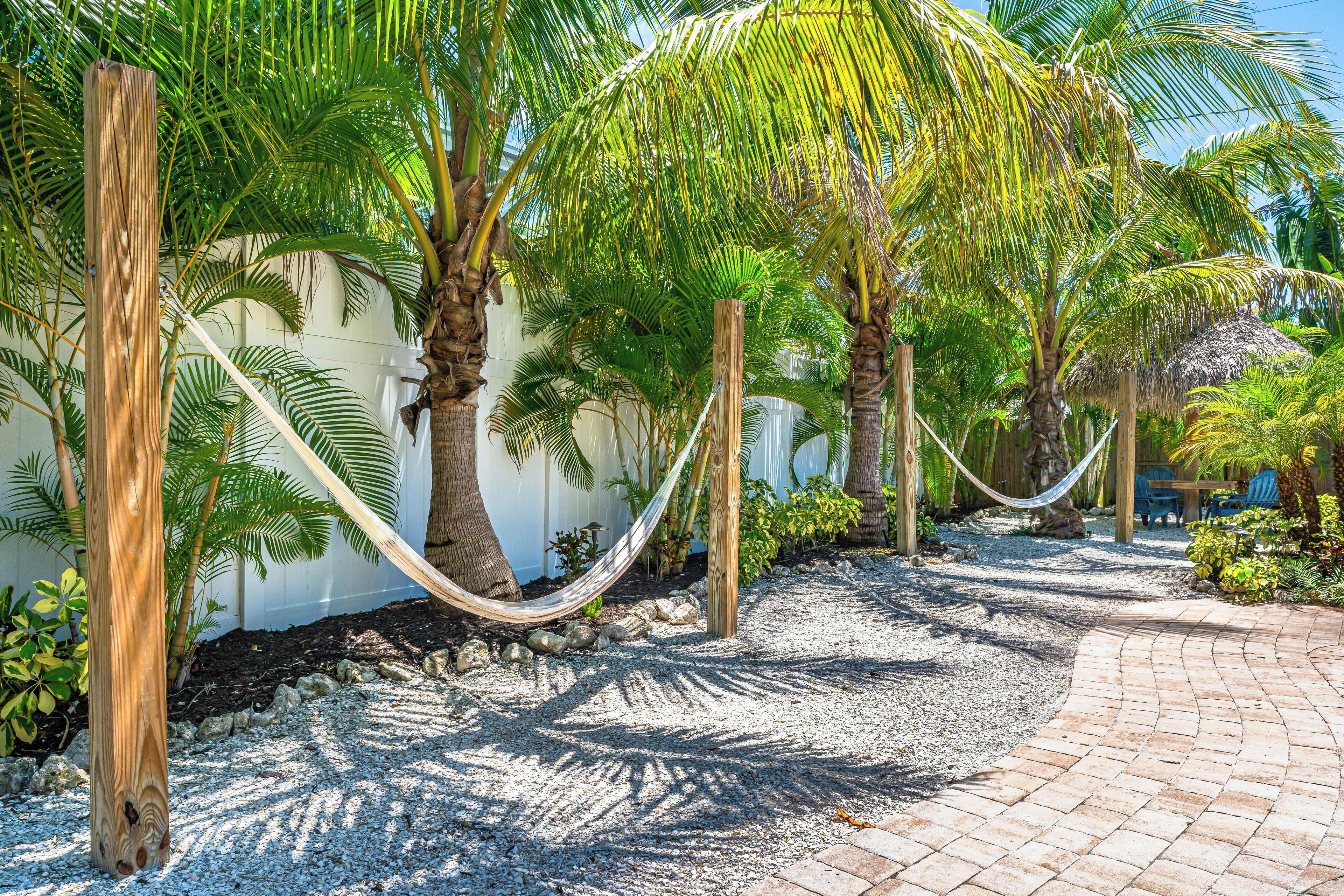A pathway lined with palm trees and a hammock, leading to a seating area with a thatched roof.