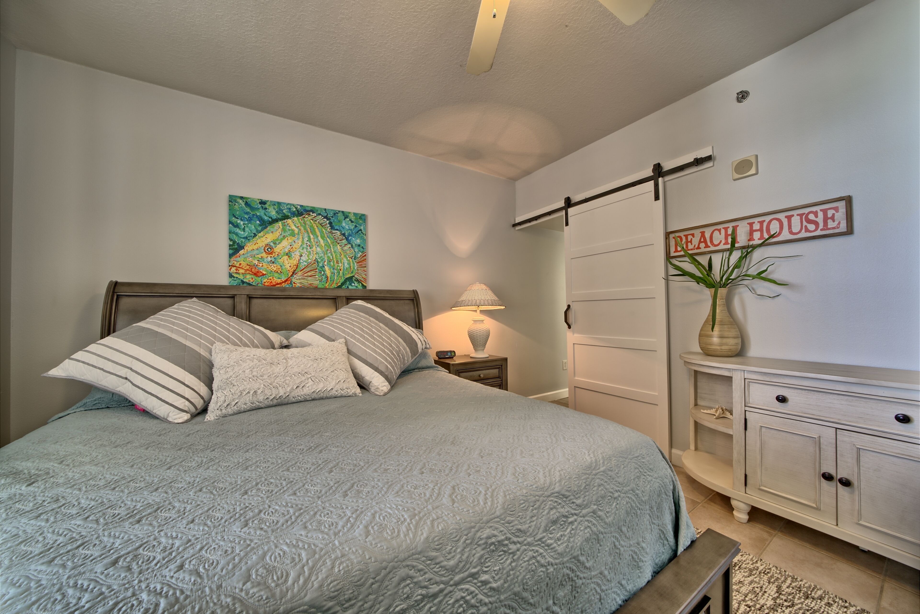 Un dormitorio con cama, mesita de noche, lámpara, cómoda, planta y un cartel en la pared que dice "Beach House".