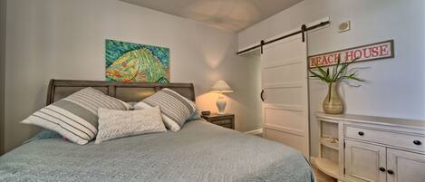 Un dormitorio con cama, mesita de noche, lámpara, cómoda, planta y un cartel en la pared que dice "Beach House".