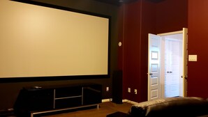 Une salle de cinéma maison dotée d’un grand écran, d’un meuble de divertissement aux couleurs sombres, d’un mur rouge et d’une porte donnant sur une autre pièce.