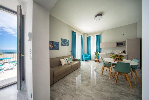 A modern living room with a sofa, dining table, and chairs, a kitchen area, and a balcony with a view of the sea.