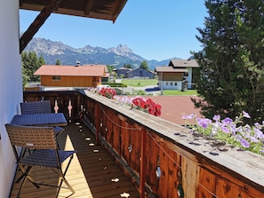 Une terrasse en bois avec une chaise en osier et une table, offrant une vue sur un paysage montagneux avec des maisons et un ciel dégagé.