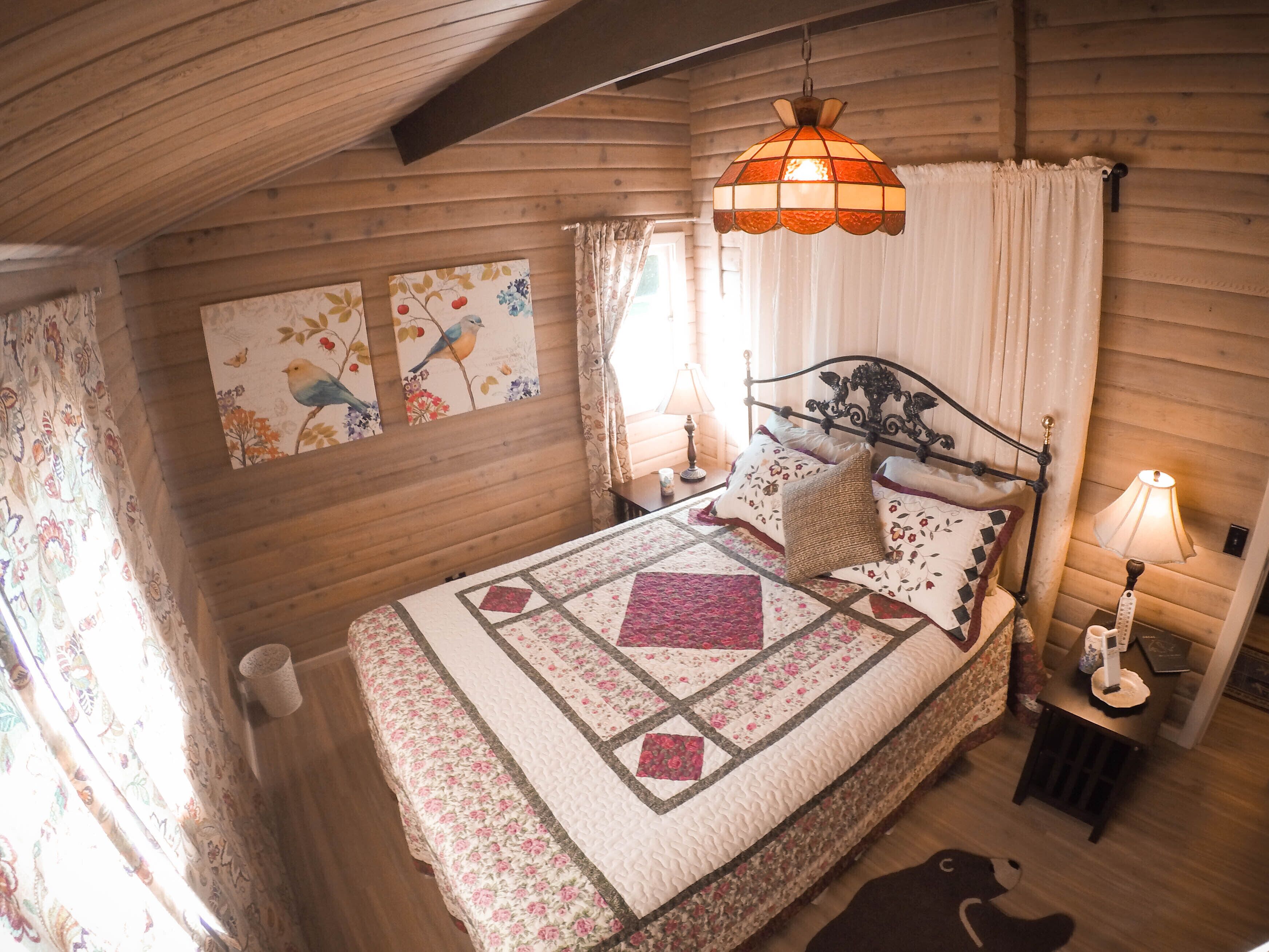 Een knus slaapkamer met een houten plafond, een bed met een sprei, een hanglamp en bloemrijke wanddecoratie.