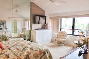 Relax in this serene bedroom with a large bed, flat-screen TV, and a view of palm trees and buildings outside.