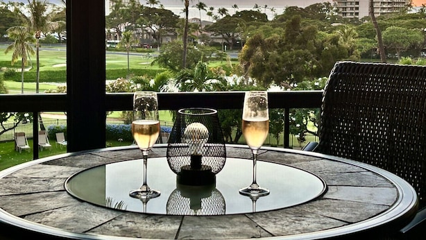 A round table with two glasses of champagne and a decorative centerpiece, overlooking a golf course and palm trees with a sunset in the background. - Family Vacation Home - Ocean Views - Walk to Beach - Sleeps 7 - Hosted by Owner (Lahaina)