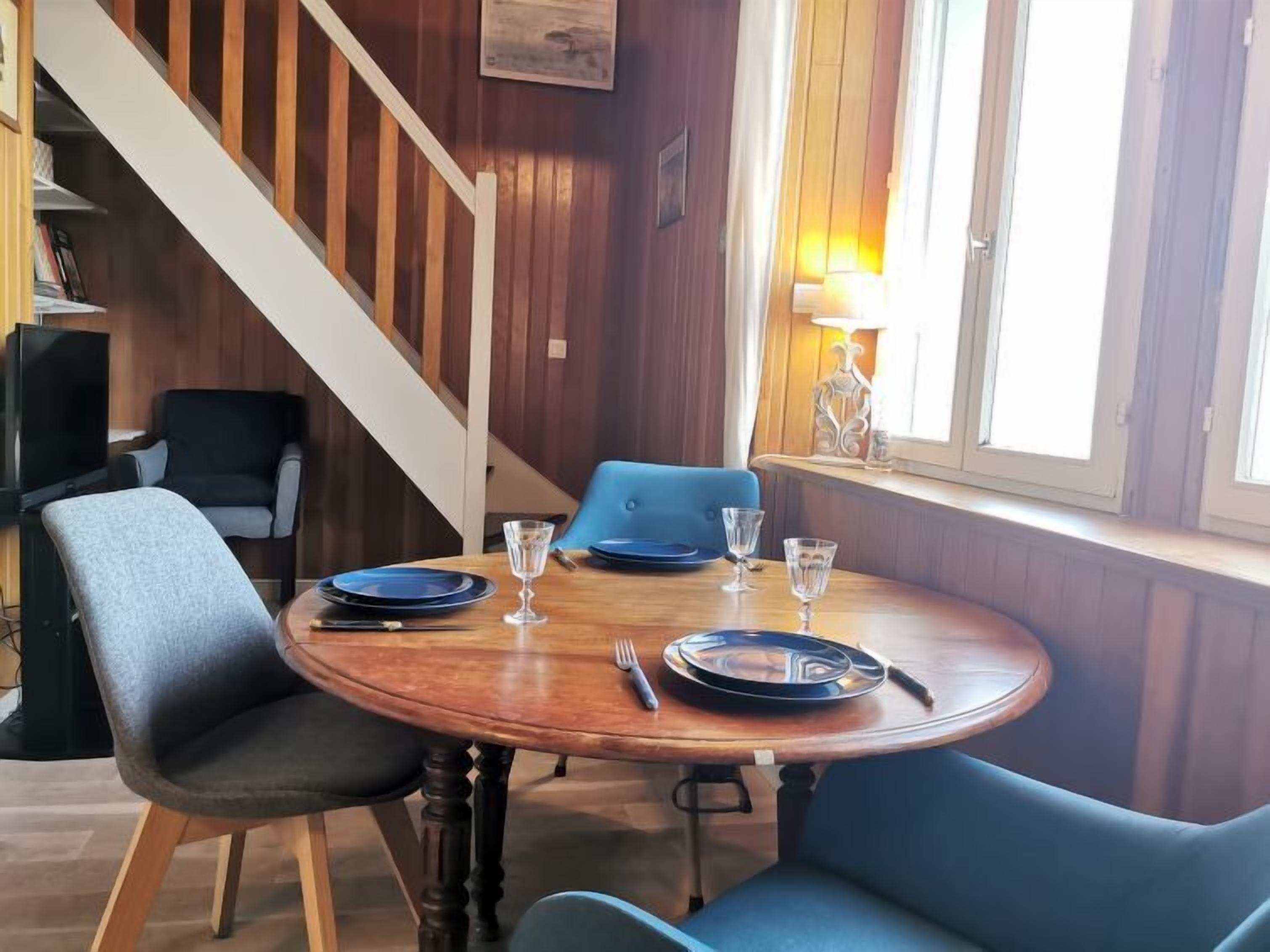 A dining area with a wooden table set for four, surrounded by blue chairs. A staircase with wooden railings is visible in the background.