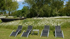 Three lounge chairs in a grassy area with white flowers, a small table, and a boat in the background. - E - Uns Schün, Germany (Middelhagen)