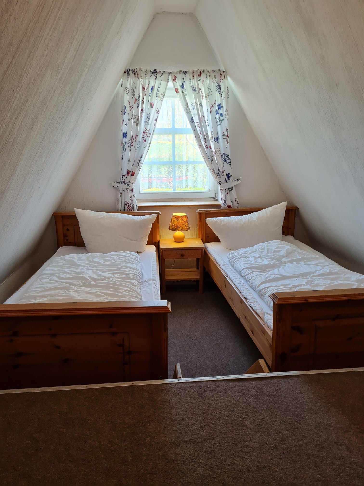 A small, cozy attic bedroom with two wooden beds, a nightstand with a lamp, and a window with floral curtains.
