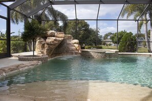 Ein Pool mit Felsformation und Whirlpool, umgeben von einer überdachten Liegefläche mit Palmen.