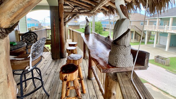 A wooden deck with a thatched roof, outdoor seating, and a view of a residential area. - LOS PELICANOS - 2 bedroom, Gulf View, Palapa Bar on deck!! (Crystal Beach)