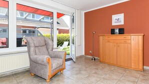 A room with a grey armchair, a wooden cabinet, and a large window with a view of a house and greenery outside. - Apartment "below" - "House Passat" (Neuharlingersiel)