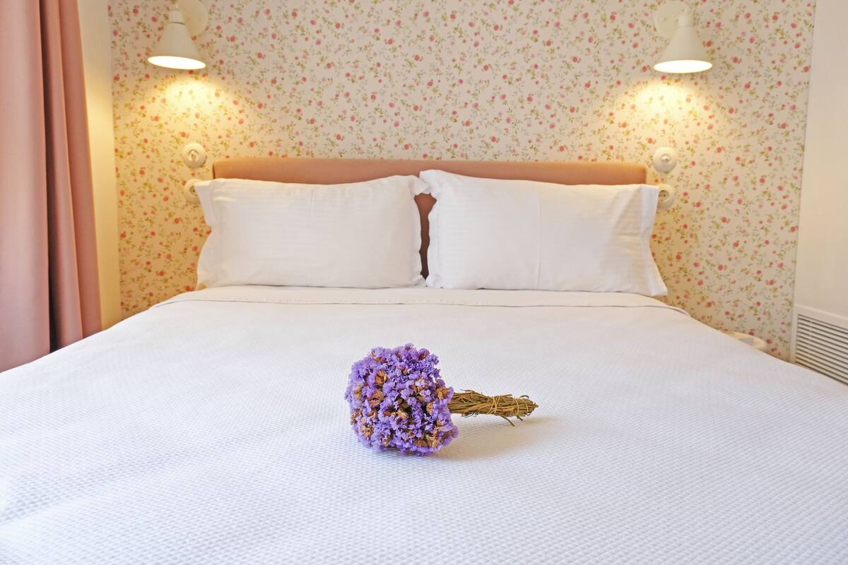 A neatly made bed with white linens and a bouquet of purple flowers on the bed.