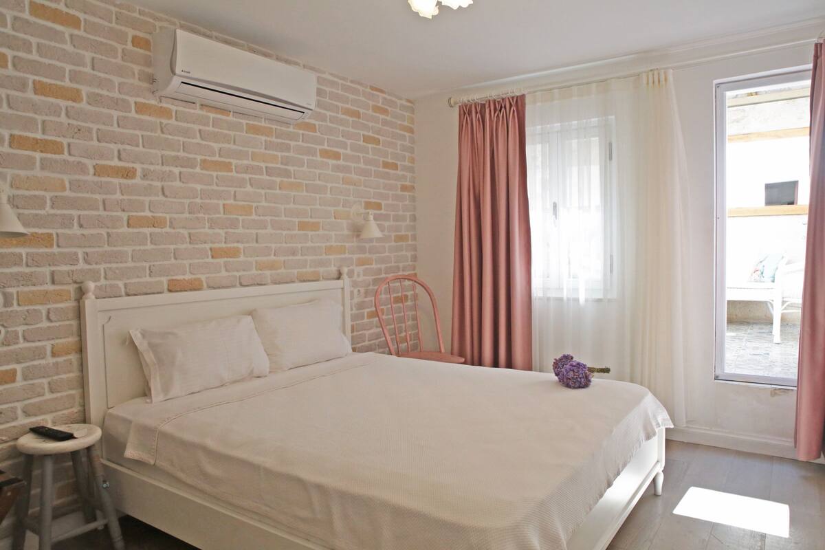 A bedroom with a brick wall, a bed, a chair, a window with curtains, and an air conditioning unit.
