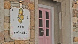 A stone building with a pink door, a sign with fruit, and a planter with flowers. - Evreka Butik Otel (Bozcaada)