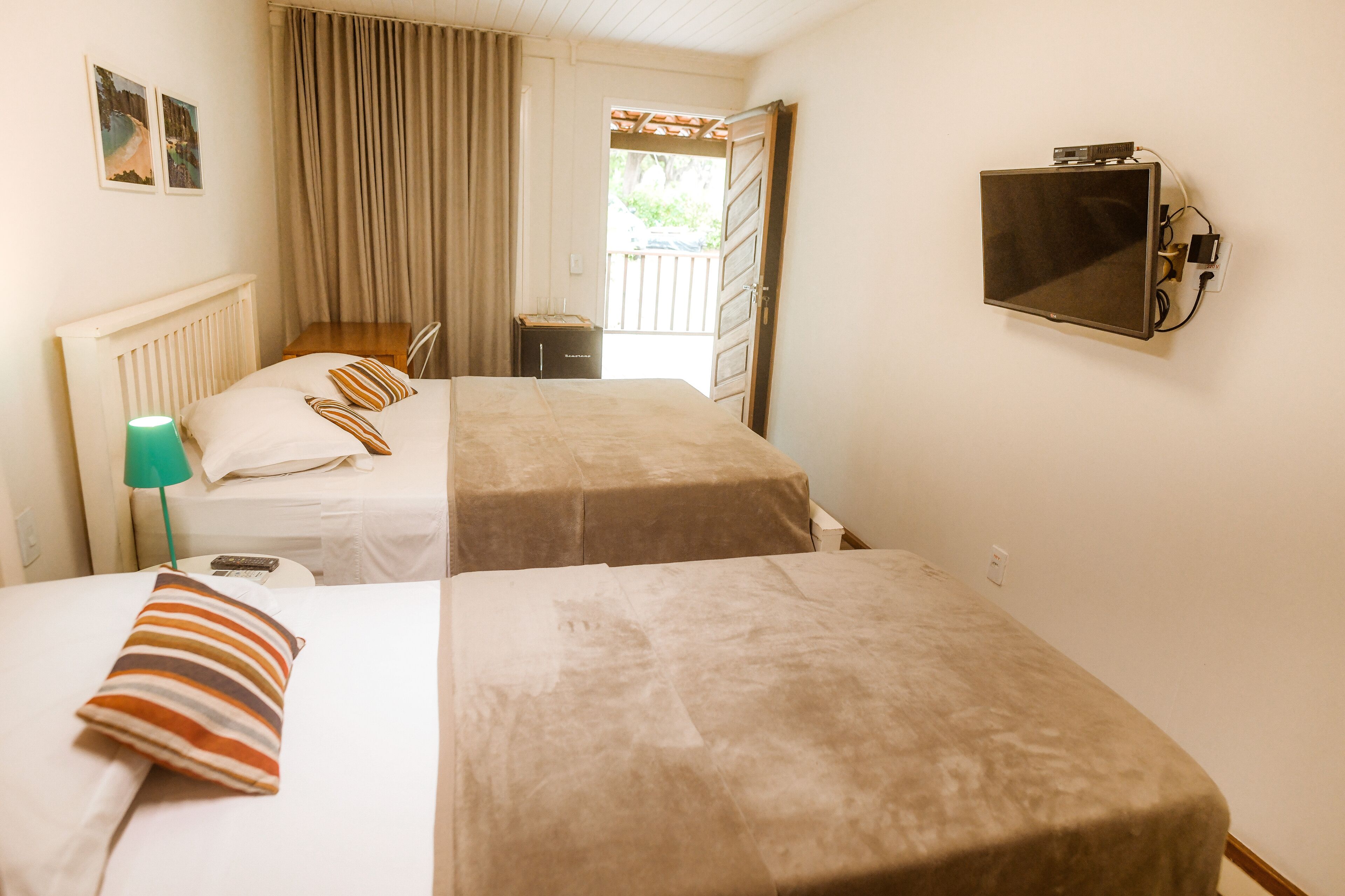 A hotel room with two beds, a TV mounted on the wall, a desk with a lamp, and a window with curtains.