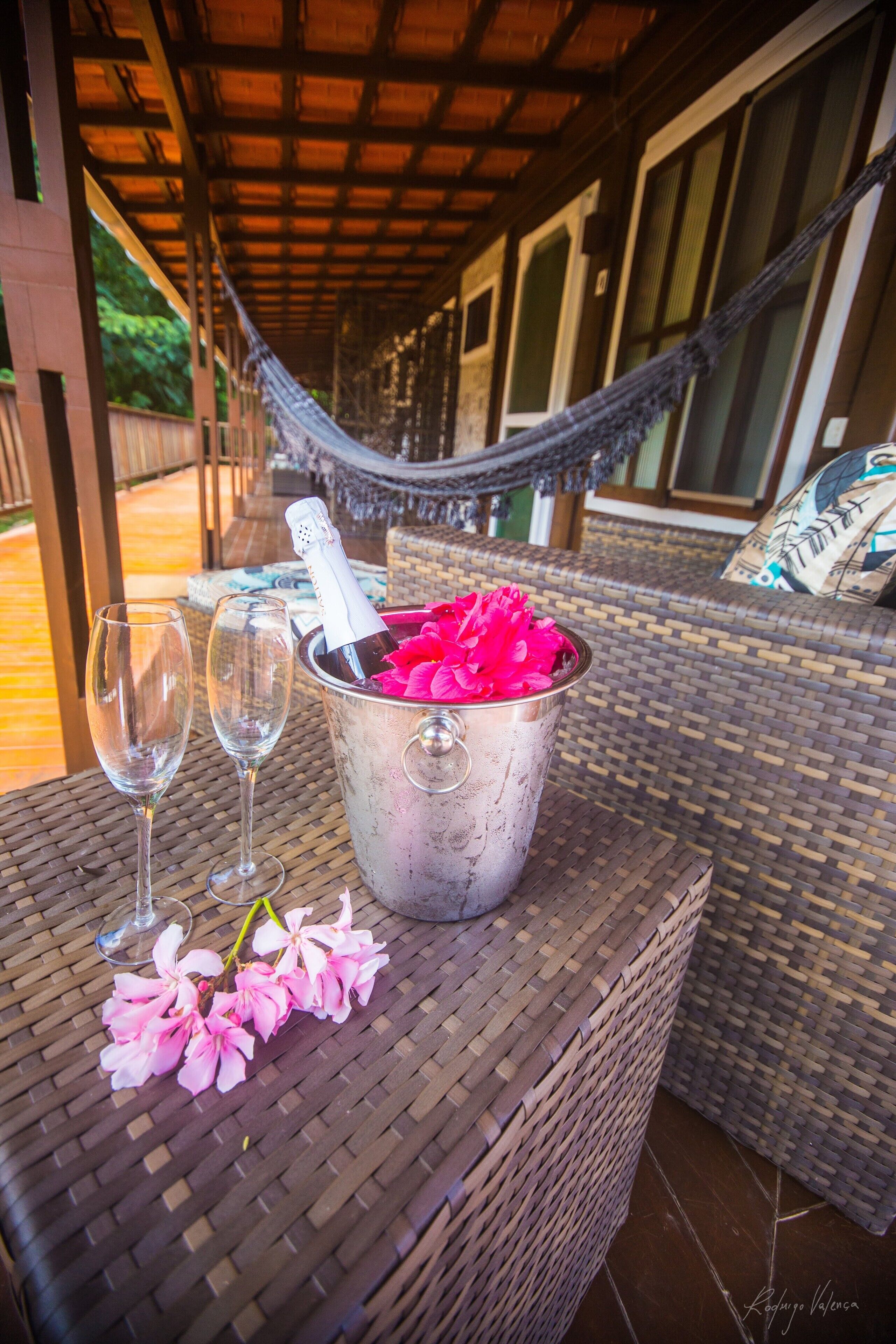A wicker table with a metal bucket containing a bottle and pink flowers, two empty wine glasses, and a hammock in the background.