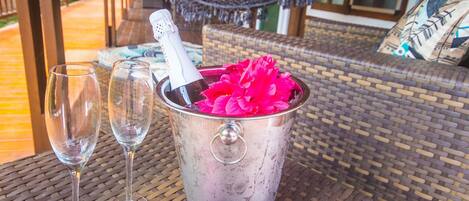 A wicker table with a metal bucket containing a bottle and pink flowers, two empty wine glasses, and a hammock in the background.