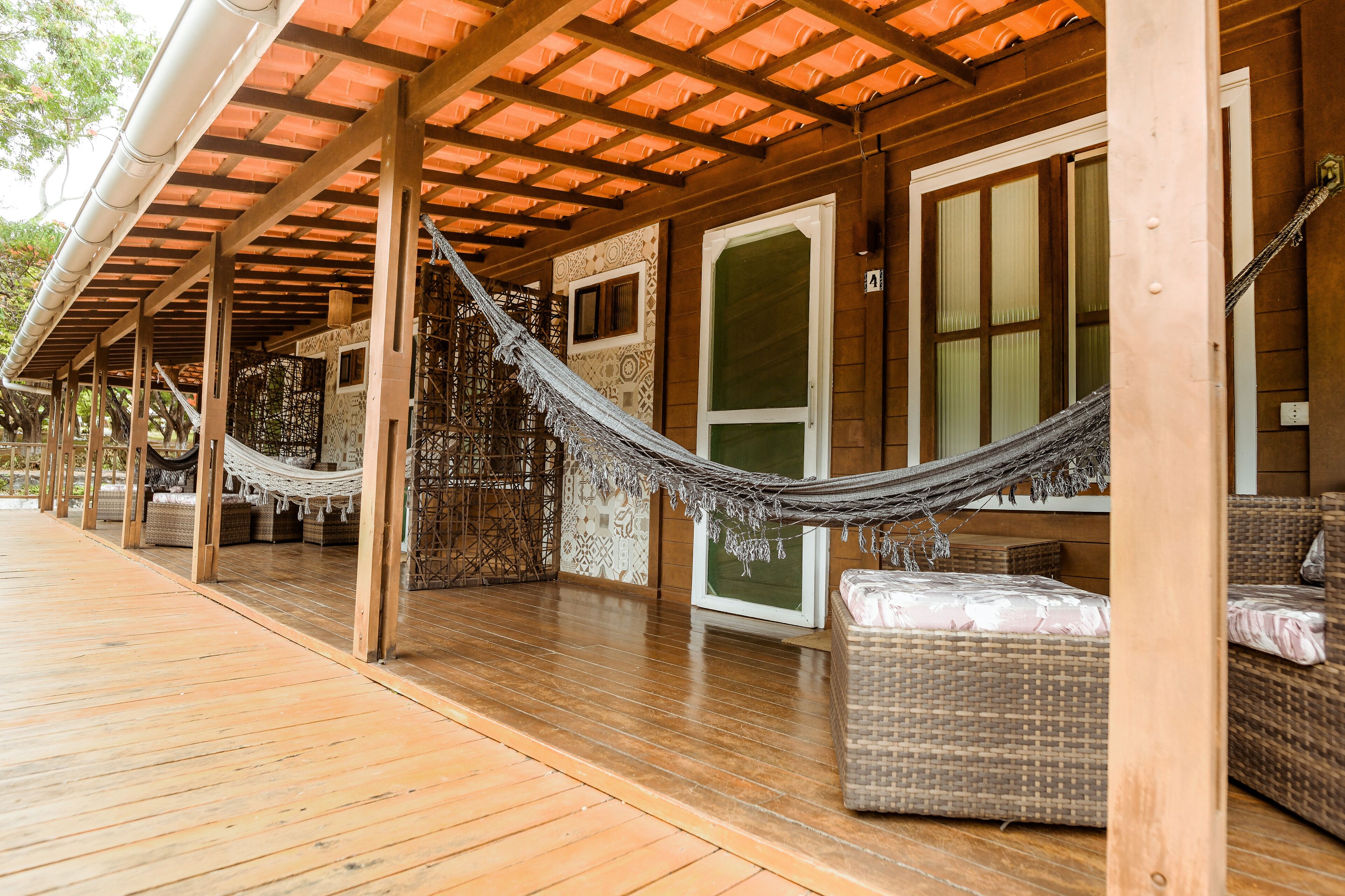 Una terraza de madera con una hamaca, mobiliario de mimbre y un área cubierta con techo de tejas.