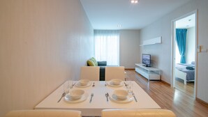 A modern dining area with a white table set for six, a sofa, a TV, and a view of a bedroom with a bed and curtains. - My Resort Beach Apartment 2 (Tambon Nong Kae)