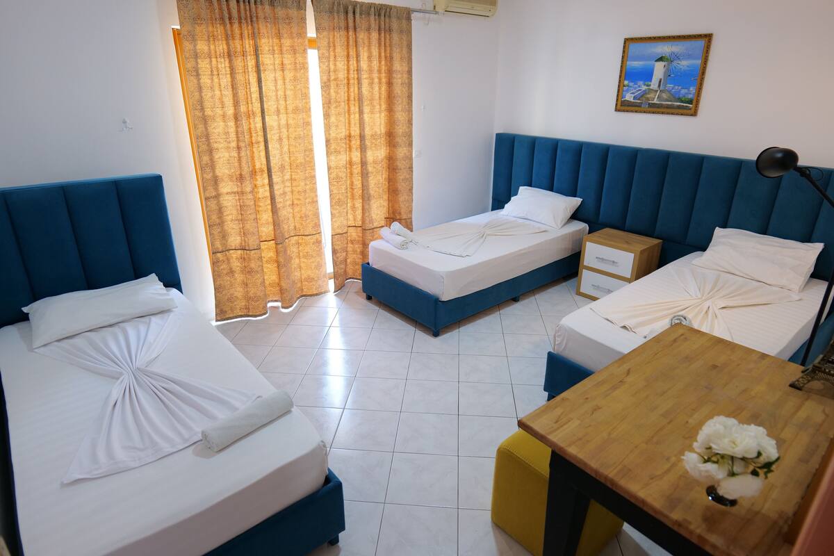 Habitación de hotel con dos camas, una mesa de madera con un florero y un cuadro en la pared, además de una ventana con cortinas.