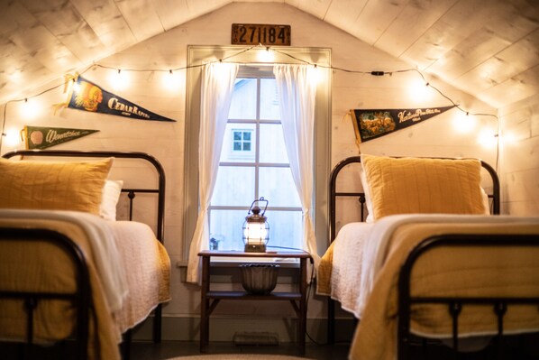 Un cuarto ático acogedor con dos camas metálicas, una mesita con un farol y una ventana con cortinas.