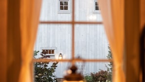 A view through a window with curtains, revealing a white building with a cross on the roof, a small table with a candle, and some greenery outside. - Historic Cottage in the Heart of the Village (Granville)