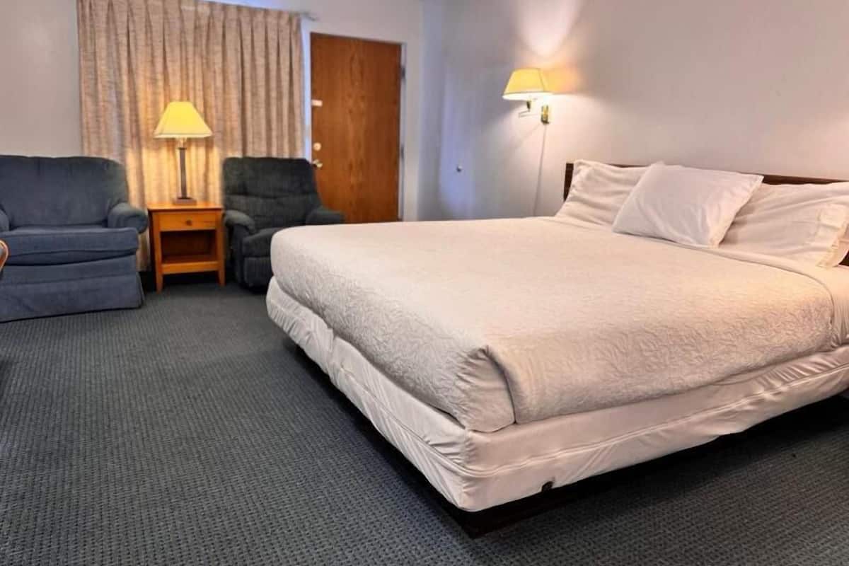 Een hotelkamer met een groot bed, twee stoelen, een houten nachtkastje, een lamp en een deur.