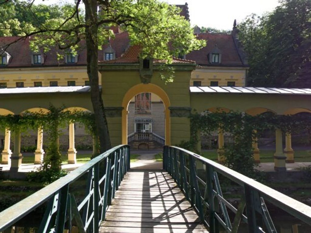 Eine Holzbrücke führt zu einem Gebäude mit einer gelben Fassade und einem roten Ziegeldach.