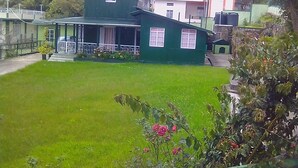 Una casa de dos pisos con techo verde y paredes blancas, rodeada por un césped cuidado y plantas florecientes.