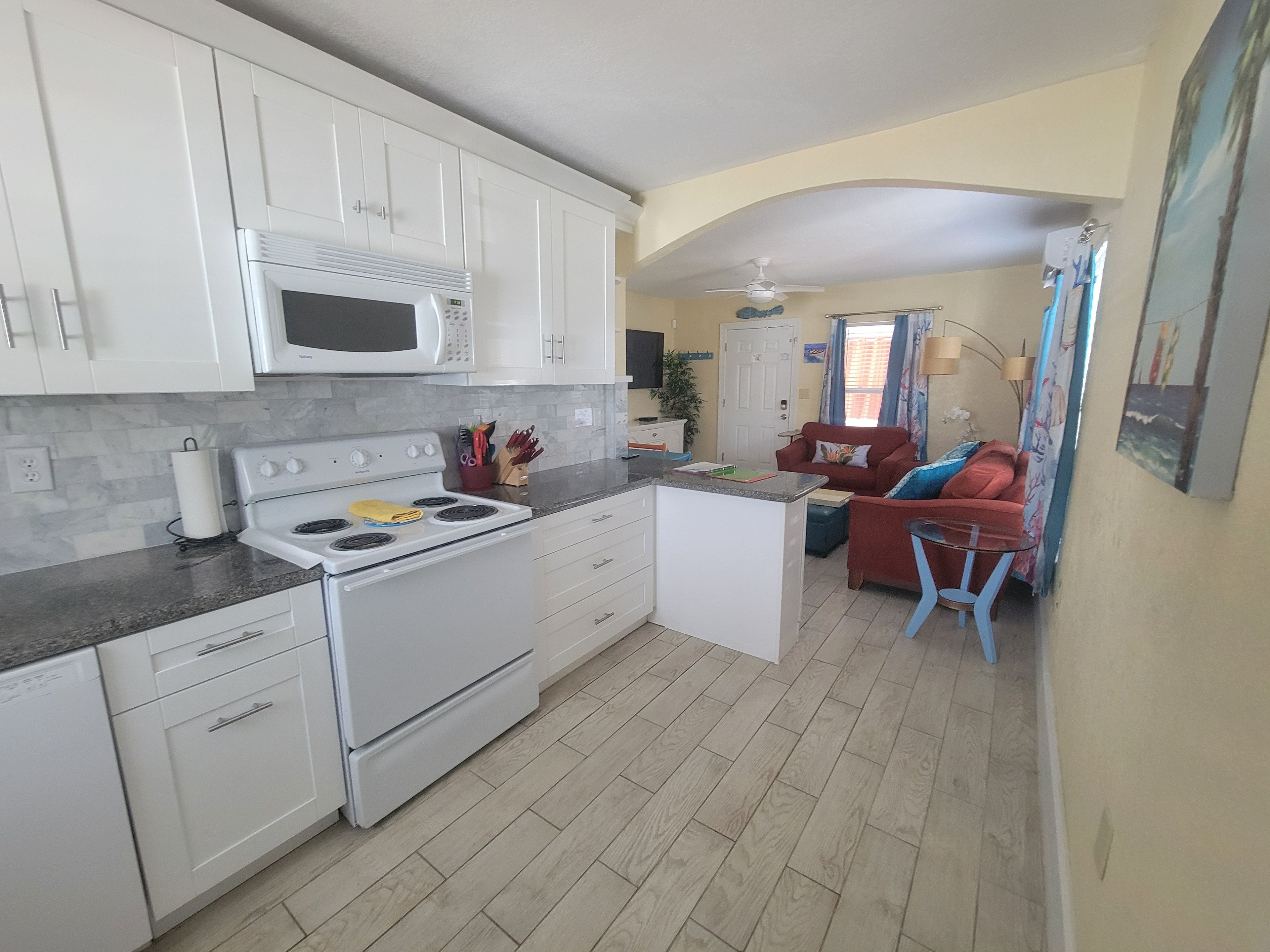 A compact kitchen with white cabinets, a microwave, and a stove. There is a small island with a sink and a red chair. The room has a tiled floor and a ceiling fan.