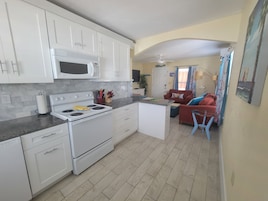 Cocina compacta con gabinetes blancos, microondas y estufa. Hay una pequeña isla con un lavamanos y una silla roja. El cuarto tiene piso de baldosas y un ventilador de techo.